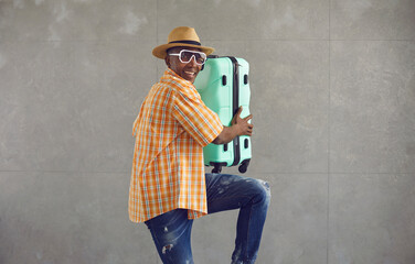 Studio portrait of happy positive young black tourist in sunglasses and sun hat carrying his travel...