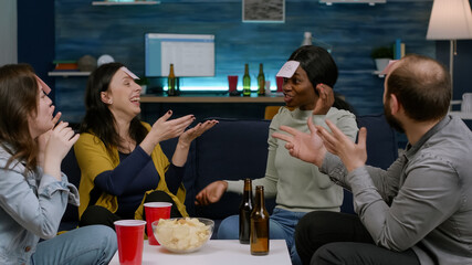 Multiracial group playing Guess who game with sticky papers attaching to foreheads. Multi-ethnic friends having fun, laughing together while sitting on sofa in living room late at night