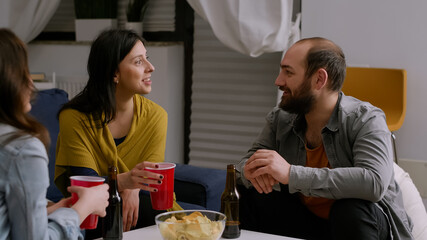 Multi-ethnic friends socializing during night home party while resting on couch in living room drinking beer having fun. Group of multi-ethnic people hanging out enjoying time spending together