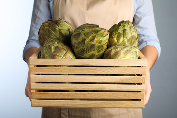 Woman holding wooden crate with fresh raw artichokes on grey background, closeup