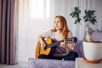 Beautiful young adult girl with long red hair playing the guitar sitting at home in light interior, selective focus. Thoughtful woman plucks the strings, composing music. Home leisure, hobbies.
