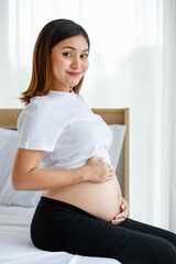 happy smiling young beautiful Asian pregnant woman sitting on a bed looking and touching her belly with care and love. Expecting for a strong and healthy baby