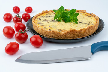 Closeup wide studio shot of freshly baked yellow French salty cake, or quiche, with mushrooms, red cherry tomatoes and green mint, on black design plate, isolated on white background. Kitchen knife.