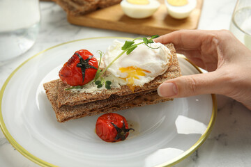 Woman eating fresh rye crispbread with poached egg, cream cheese and grilled tomato at table, closeup