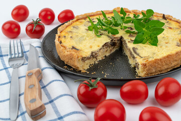 Closeup wide studio shot of freshly baked yellow French salty cake, or quiche with mushrooms, red cherry tomatoes, green mint and kitchen cloth, on black design plate on white background. Cutlery set.