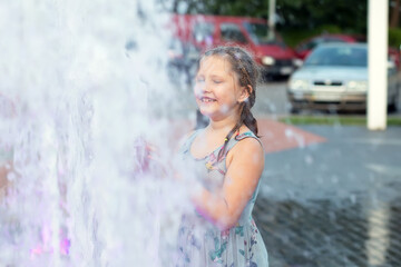 Obraz premium Little cute funny girl child bathes on a hot summer day in a public city fountain. The child runs, plays among the jets and splashes of water