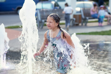 Little cute funny girl child bathes on a hot summer day in a public city fountain. The child runs, plays among the jets and splashes of water