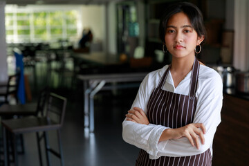 Confident ethnic female waitress in apron standing with crossed arms in cafe and looking at camera