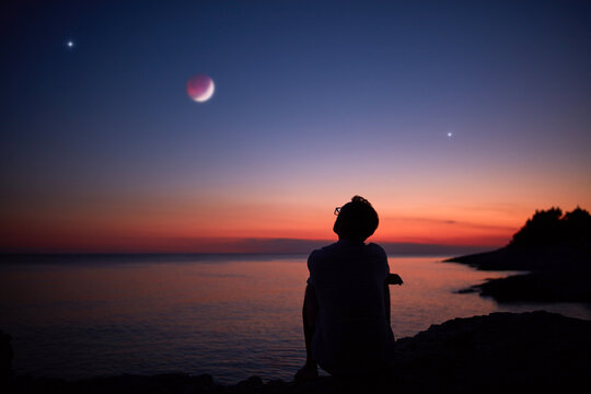 Silhouette of a man looking at the Moon and stars over sea ocean horizon.