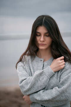 Teanager with long dark hair on the beach during cloudy evening. Brunette girl wears clothes near the sea during cold evening. Isolated portrait of lonely woman.