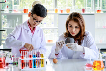 Young Asian woman with magnifier examining chicken egg while focused boy mixing colorful liquids during scientific experiment in chemistry laboratory