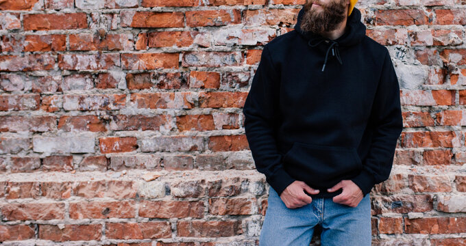 City Portrait Of Handsome Guy Wearing Black Blank Hoodie And Baseball Cap With Space For Your Logo Or Design. Mockup For Print