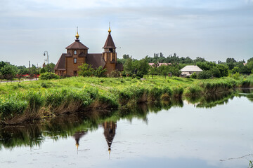 Obraz premium An old wooden church on the bank of the river. Reflections can be seen in calm water and residential buildings in the distance. 