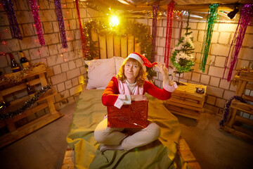 Nice girl in a red dress and hat of Santa with big red gift box in room decorated for Christmas. Woman in beautirul studio during photo shoot before New Year. Female model posing in holiday.