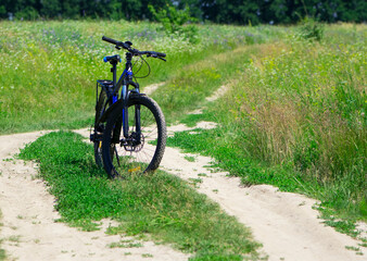 bike stands on the road in the field. A mountain bike stands on a field path with green grass. Mountain bike, blooming summer field, meadow flowers, sunny day. ride a bike.