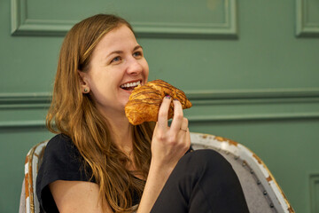 Beautiful woman with croissant on a green background. Caucasian adult girl indoors laughing and eating. Modern fast paced and take away snack.