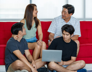Fototapeta premium Four members Asian family consisting of parents, two adult sons happily spending time together during holidays, using laptop in comfortable living room at home. Presenting modern technology concept.