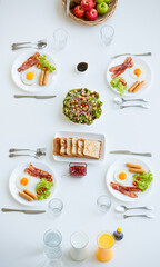 Top, aerial view of four breakfasts served on homey clean white table consists of fried eggs, bacon, sausages on  plates with spoons, forks and knives decorated by pieces bread, healthy salad, apples