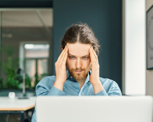 Concentrated male company employee with fingers on temples and looking at laptop computer screen, trying to concentrate while sitting at workplace in office. Young man suffering from headache at work
