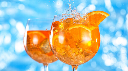 Ice cube falling into glass of orange cocktail. Aperol spritz with sea surface background. Close-up.