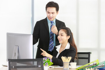 Asian woman sitting on a desk pointing at a desktop computer and a Businessman standing glad at work in the office. Concept meeting team for woirking.