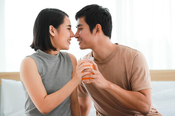 Portrait shot of cute smiling young Asian lover couple sitting on a bed together at home in the morning. Wife holding and drinking a glass of orange juice with her husband whispering her a secret