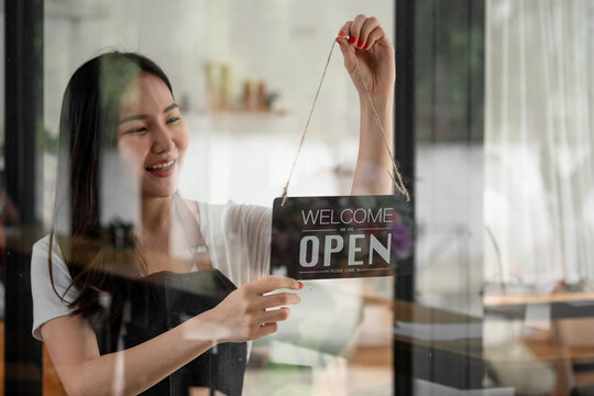 Portrait of smiling young barista girl in apron holding open sign board while standing at her cafe. elegant asian coffee shop female staff turn door plate in the morning in own store small business