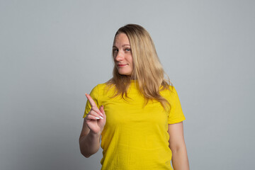 Beautiful mature woman smiling and pointing finger upward, wearing in casual yellow t shirt, gray background