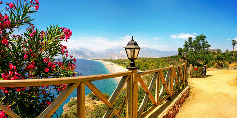 Panorama resort Antalya with soft focusing in background. Sea, wide beach, blue sky, white clouds and chain of mountains. Scarlet oleander bush, wood fence and retro-style lanterns add charm to place.