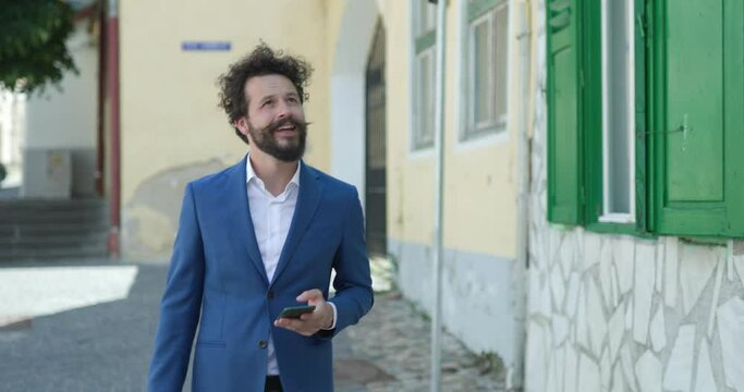 sexy bearded guy with long moustache and blue jacket holding suitcase and mobile, checking social, media, looking around and smiling while walking in medieval town from Romania