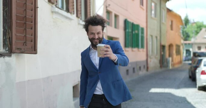 happy young man walking in medieval city, holding suitcase, drinking coffee and having a good time, smiling
