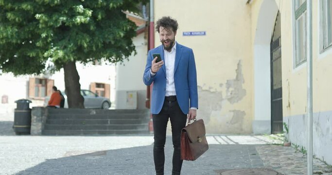 smart casual fashion model with long beard holding mobile and suitcase, reading messages and getting great news, joyful jumping and dancing, celebrating and being happy outside in the city