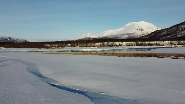 flying over snow patch covered river shore with clean cold melting water running through the river and mighty forest and snowy peak mountain in the backdrop, aerial video