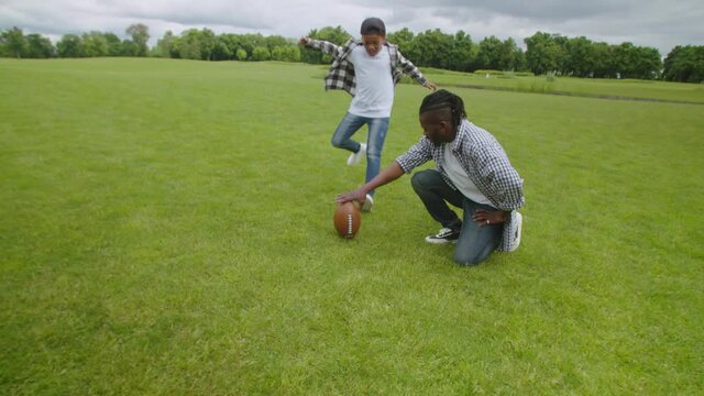 Caring handsome african father with dreadlocks teaching cute school age son playing american football, setting ball for place kick and boy kicking field goal while playing football on field.