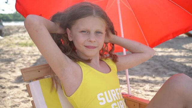 Young pretty girl in yellow summer bikini laying on beach chair with red umbrella and touching her hair.