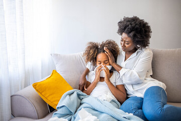 Mother helping daughter blowing her nose on the sofa at home. Ill African American girl blowing a...