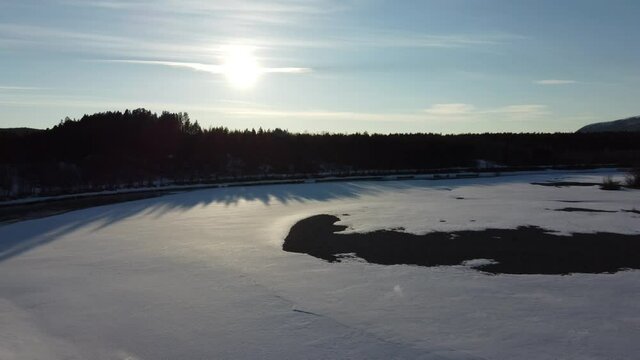 Flying over flowing river current and white snow covered beach towards the dawn sunset in early spring, aerial shot