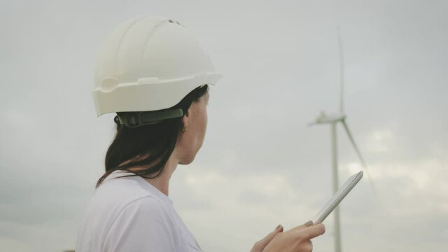 Ecology electrical energy. Woman engineer in helmet works with a tablet computer on the background of wind turbine power generator station.