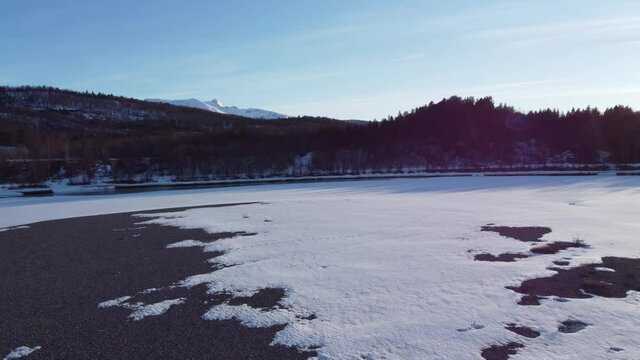 flying over snow patch covered river shore with clean cold melting water running through the river and mighty forest and snowy peak mountain in the backdrop, aerial video