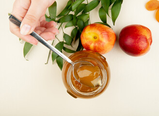 top view of female hand holding a spoon with peach jam over the glass jar and fresh nectarines with green leaves on white background