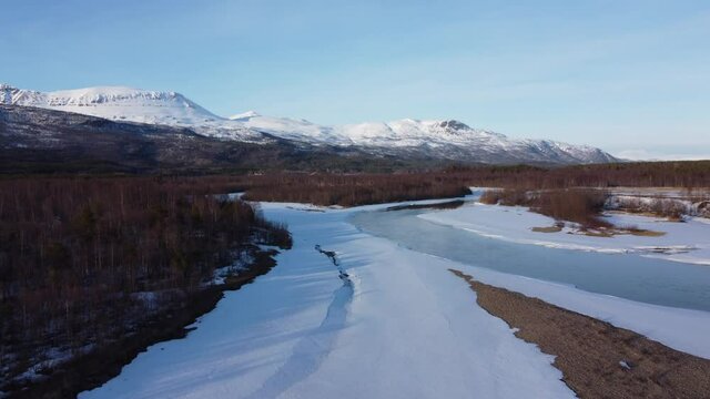 flying over snow patch covered river shore with clean cold melting water running through the river and mighty forest and snowy peak mountain in the backdrop, aerial video
