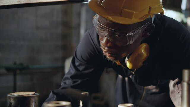 American industrial black young worker man with yellow helmet and ear protection surveillance for examination in front machine, Engineer inspection checking work in the industry factory. slow motion