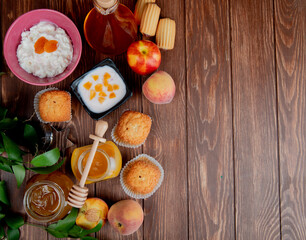 top view of peach yogurt in a black bowl and fresh ripe peaches with muffins cookies and cottage cheese with dried apricots in a bowl and glass jar with honey on wooden rustic background with copy sp