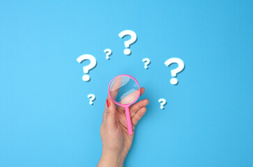 female hand holds a plastic magnifying glass and question marks on a blue background