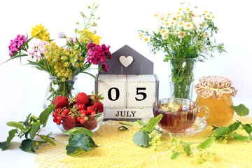Calendar for July 5 : the name of the month of July in English, cubes with the numbers 0 and 5, bouquets of wild flowers, jam, fruit, a cup of tea on a yellow openwork napkin