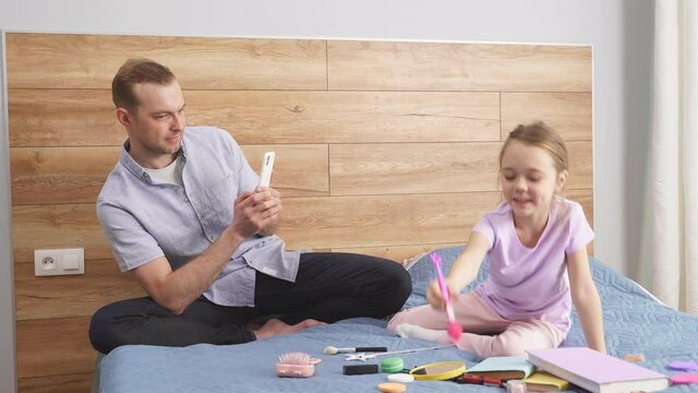 Trendy father take photo of his fashionable daughter using cosmetics, man and child sit on bed together, have fun, full of joy and happiness. Family concept.