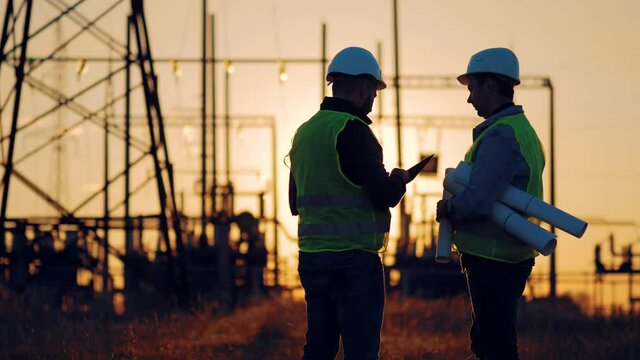 Teamwork concept. Silhouette engineer and technician work together to accomplish this task. Electrical engineers working on electricity pylon high tension power line repairs and maintenance at sunset.