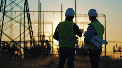 Silhouette of engineers looks at the construction of high-voltage power. Teams engineer looking discussing plan. Two engineer standing on field with electricity towers at sunset.