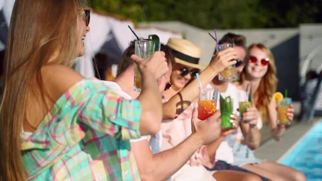Group of friends having fun at poolside party clinking glasses with fresh cocktails sitting by swimming pool on sunny summer day. Woman toasting, drinking beverage of plastic straw at luxury villa.