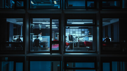 Shot from Outside the Window: Businessmen and Businesswomen Working in the Office. Managers and Specialists Doing Financial Business in the Evening. Employees Work on Computers and Delegate Tasks.